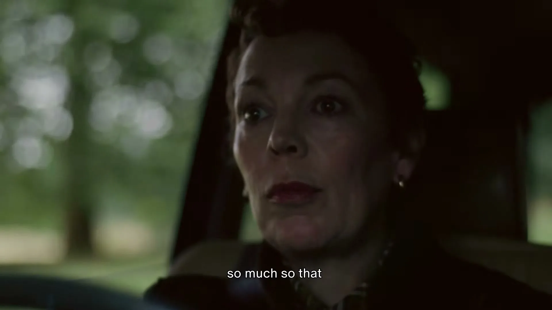 Olivia Colman, a woman in her 50s, drives a car, talking and looking around, with trees visible outside the window.