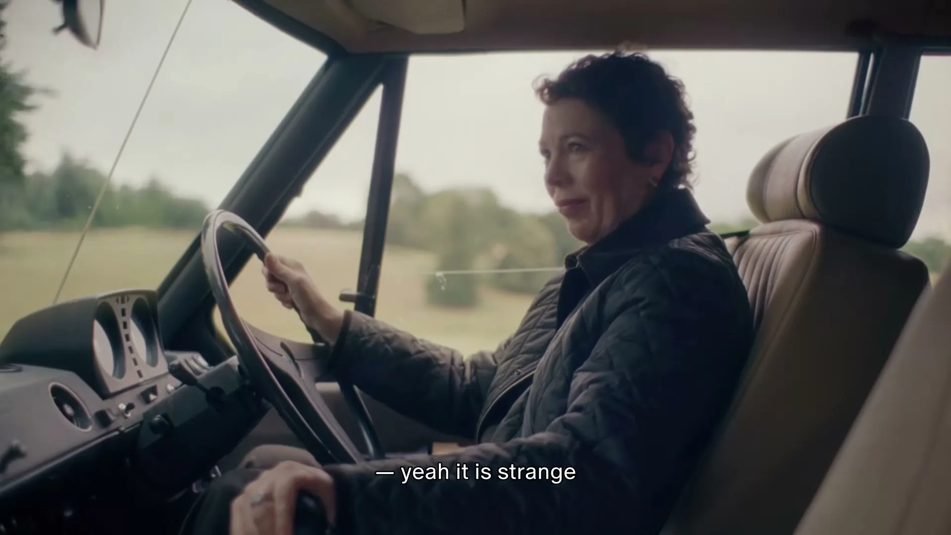 Olivia Colman drives a vintage car through a rural landscape, looking around and talking, wearing a quilted jacket.