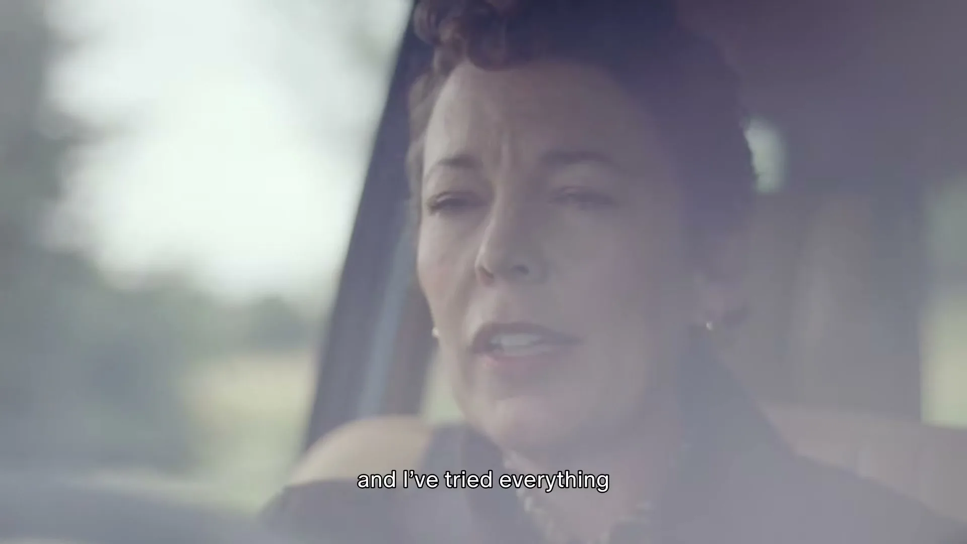 A woman with short, curly brown hair drives a car, looking forward and speaking, with a blurred reflection of the outside visible on the windshield.