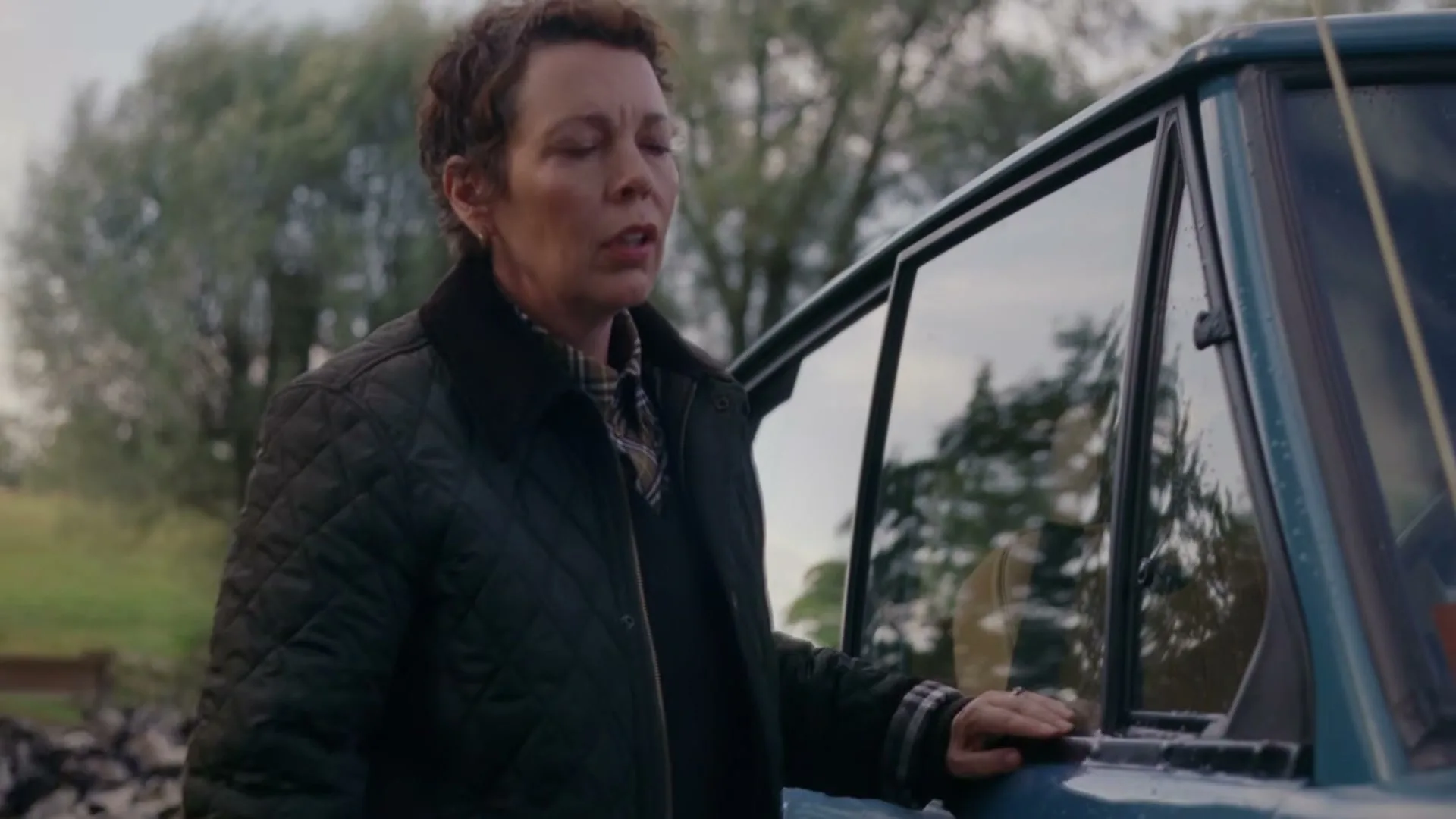 Olivia Colman, a woman in her 40s, stands next to a blue car, looking around with a concerned expression in a rural outdoor setting.