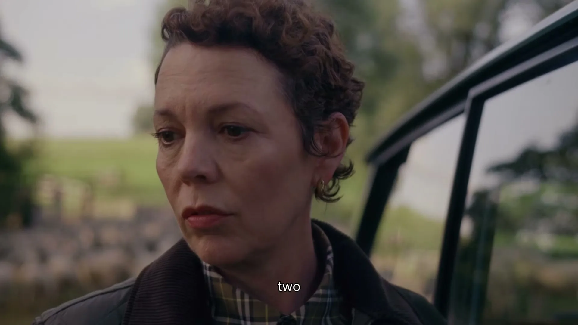 Olivia Colman, a woman with short curly brown hair, stands next to a car, looking off-camera with a thoughtful expression in a rural setting.