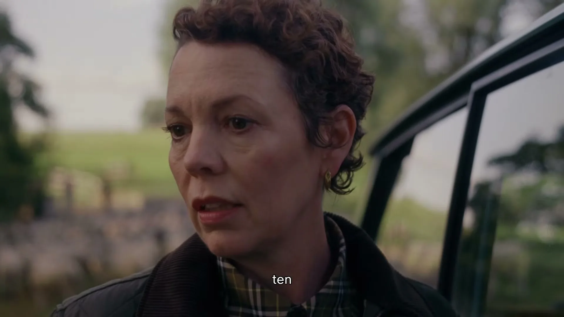 Olivia Colman, a woman with short curly hair, stands next to a car door, looking slightly concerned in a rural outdoor setting.