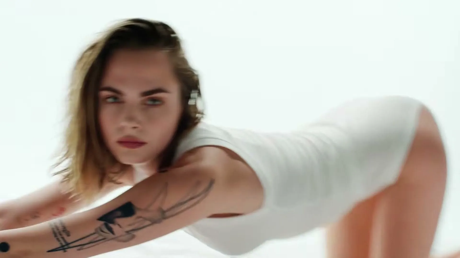 A woman with short brown hair and tattoos on her arm, wearing a white tank top, crawls towards the camera on a white surface in a studio.