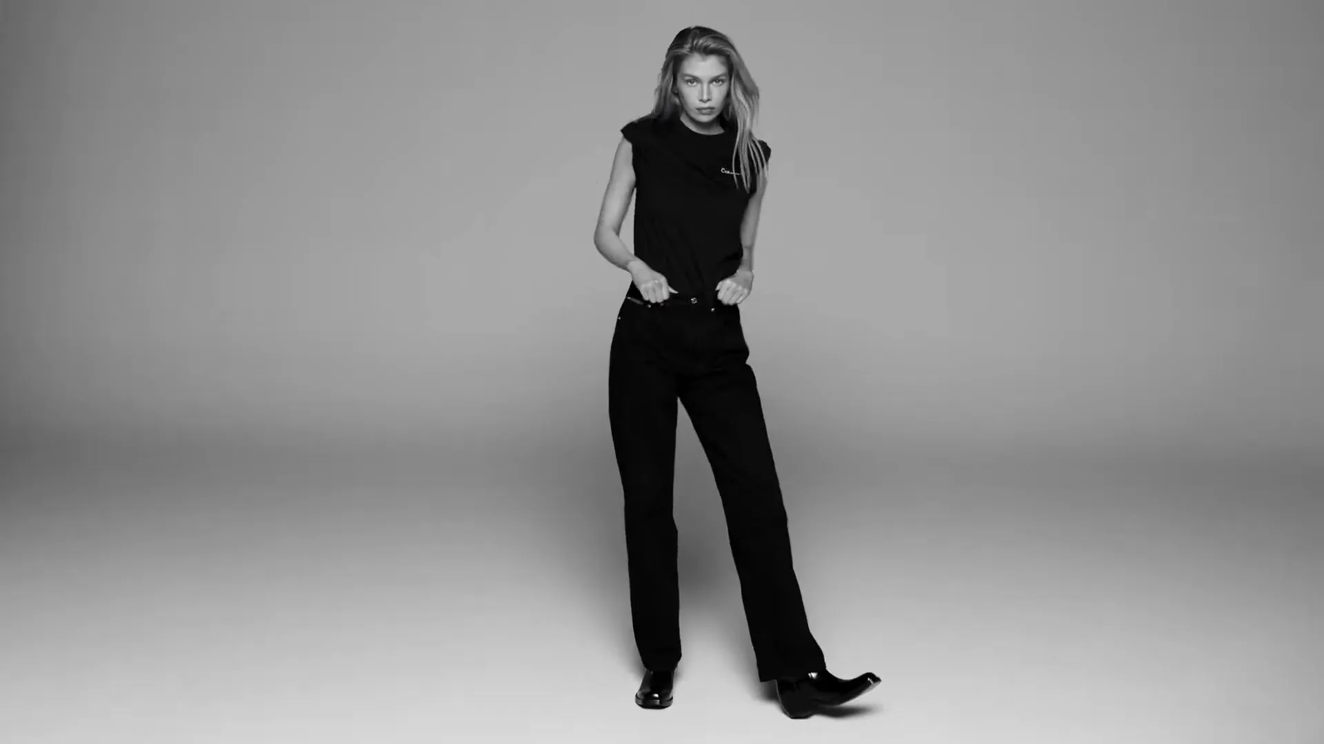A young woman with long blonde hair, wearing a black t-shirt and black pants, poses confidently in a studio.