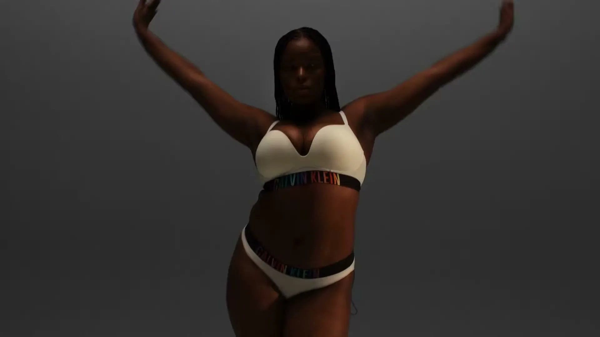 A Black woman with braids, wearing white Calvin Klein underwear with rainbow waistbands, poses with arms outstretched in a studio.