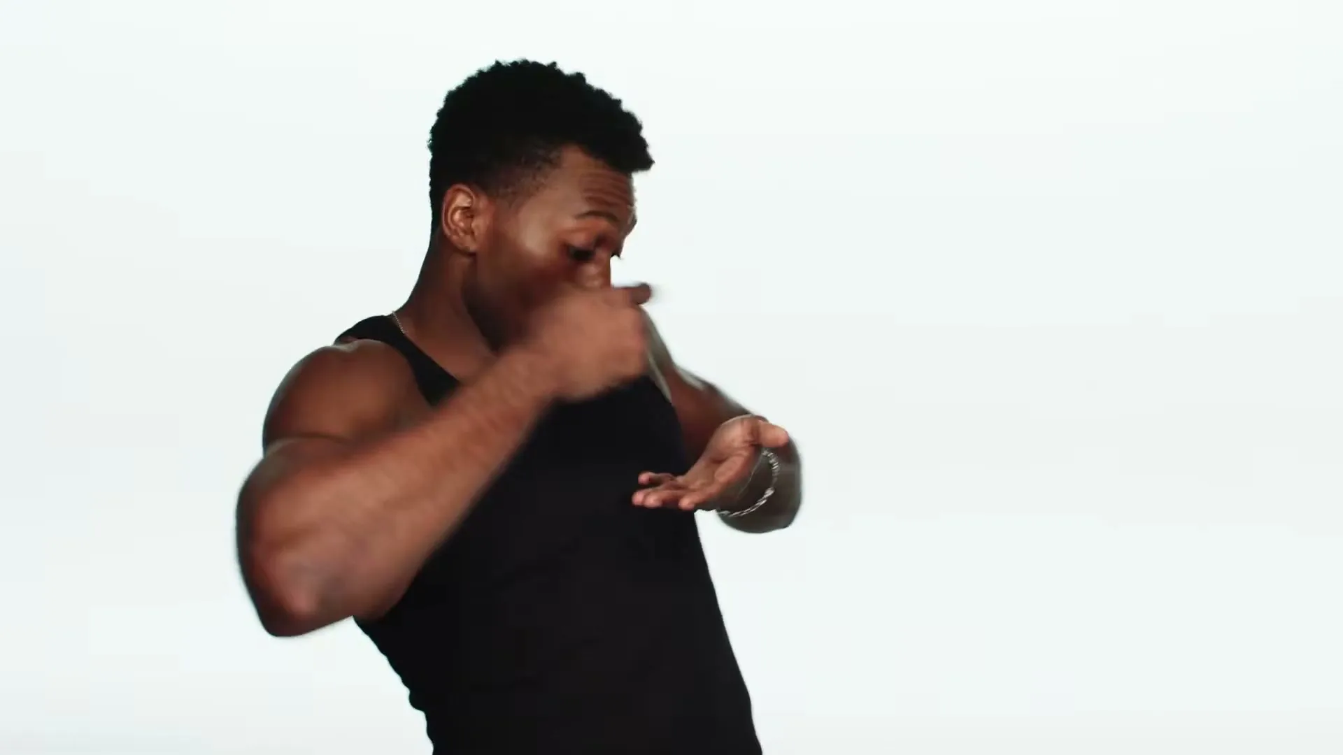 A muscular Black man in a black tank top dances energetically, making hand gestures and looking off-camera in a studio.