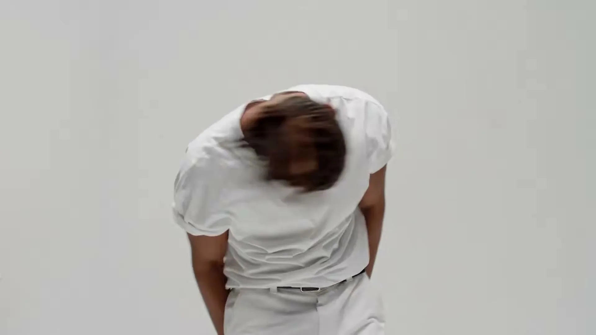 A man in his 20s, wearing a white t-shirt and white pants, dances energetically in a bright studio.