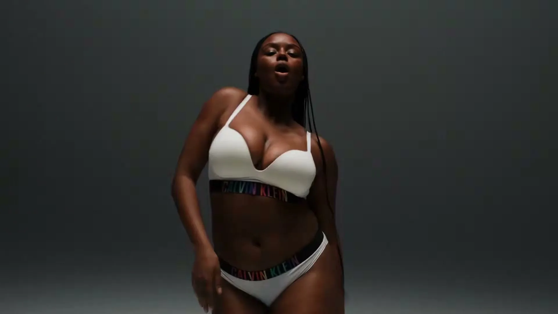A Black woman with long braids, wearing white Calvin Klein underwear, dances sensually in a studio against a gray background.