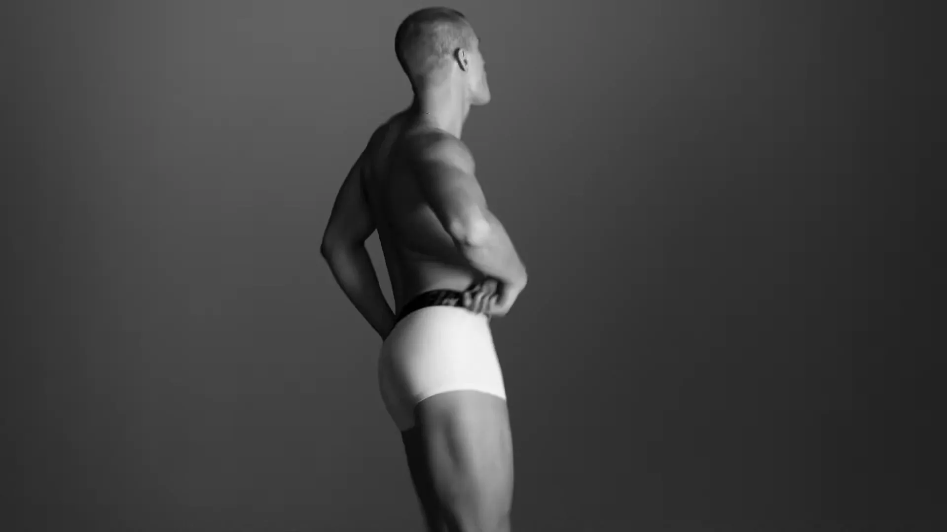 A shirtless male model in white underwear poses, turning his back to the camera in a black and white studio setting.