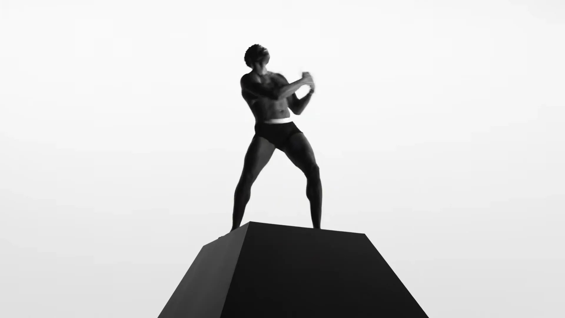 A shirtless Black man in underwear stands on a black pyramid, looking up and flexing his arms in a studio void.