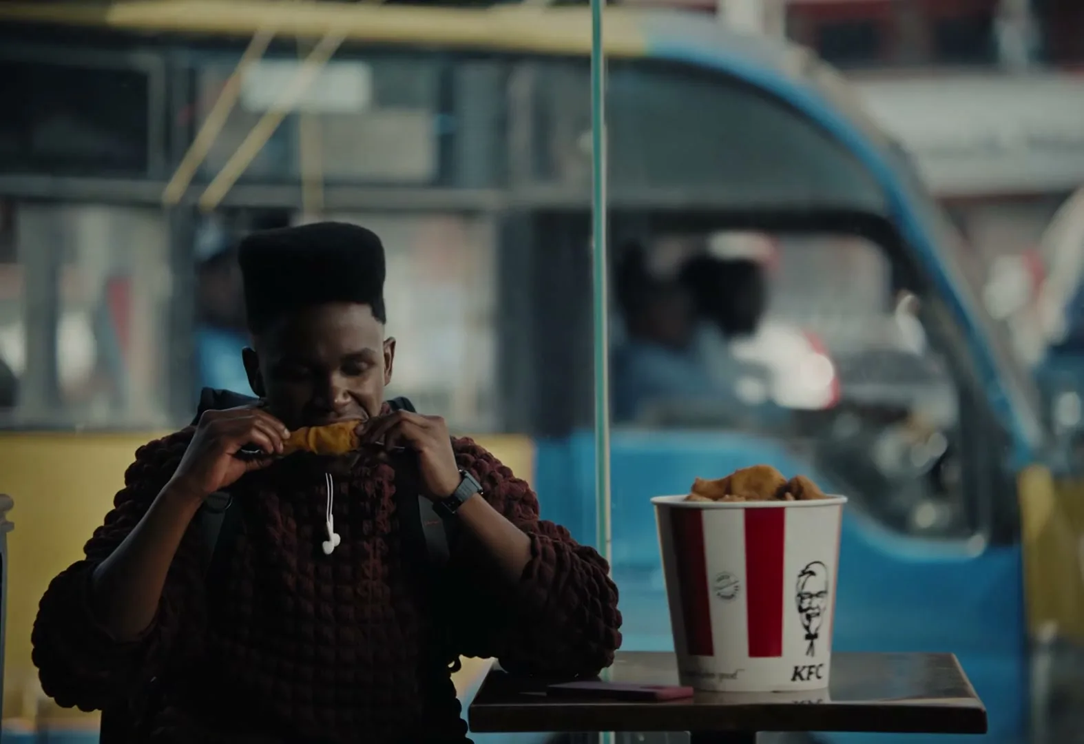 A young man with a high-top fade hairstyle sits at a table, eating fried chicken from a KFC bucket, with a busy street visible through the window behind him.
