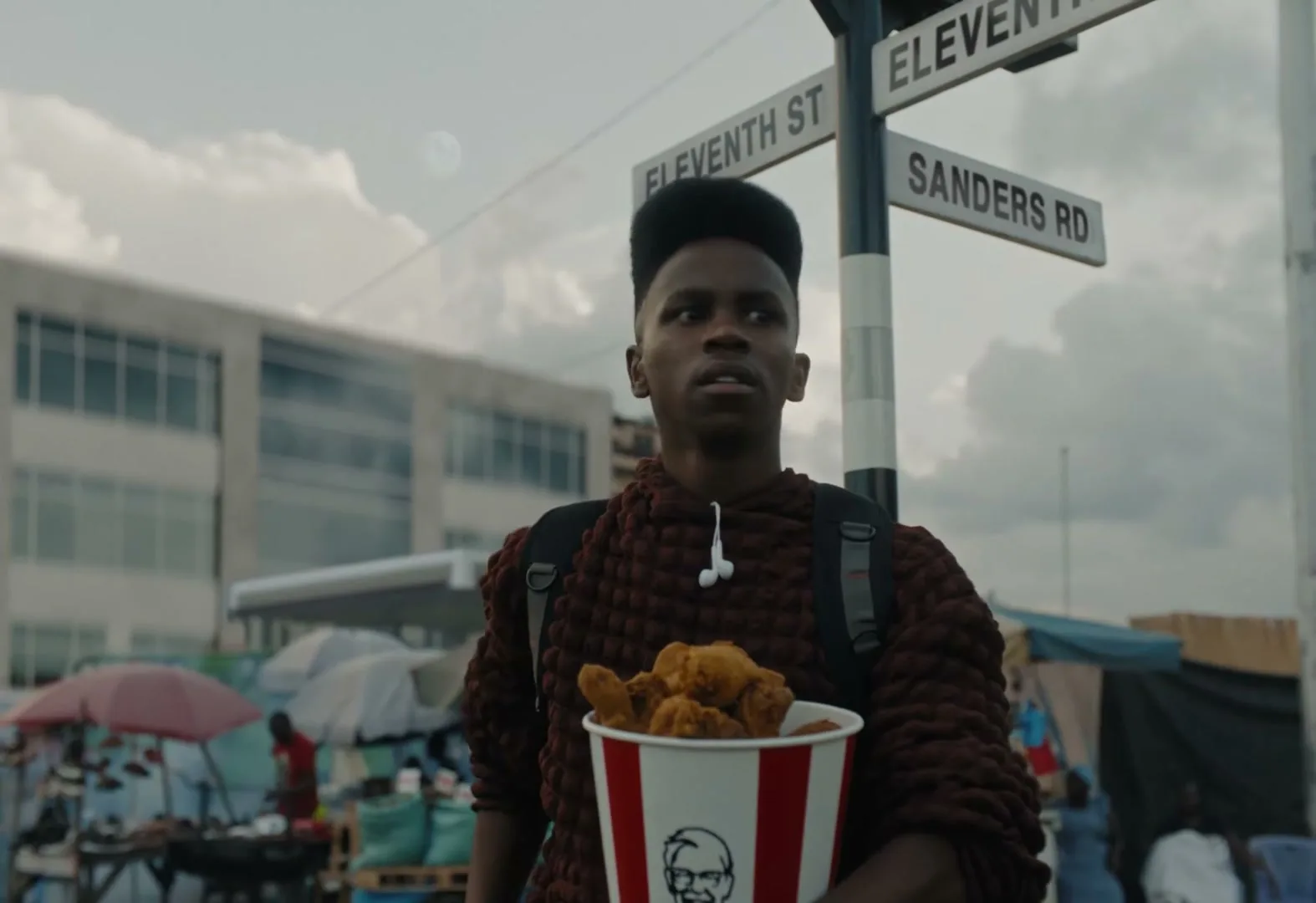 A young man with a high-top fade haircut, wearing a textured brown hoodie and backpack, walks down a street holding a KFC bucket.