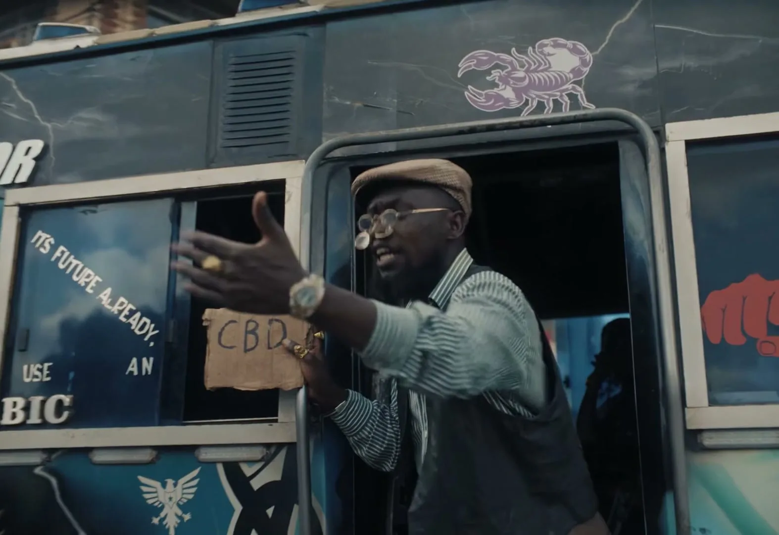 A man in a cap and glasses leans out of a bus door, gesturing with his hand and holding a "CBD" sign.