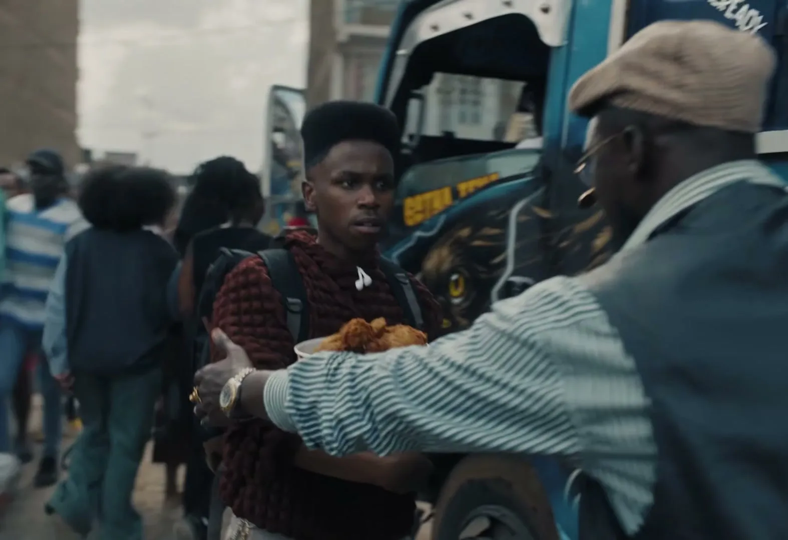 A young man with a bucket of KFC chicken is pushed by an older man in a crowded street next to a blue truck.