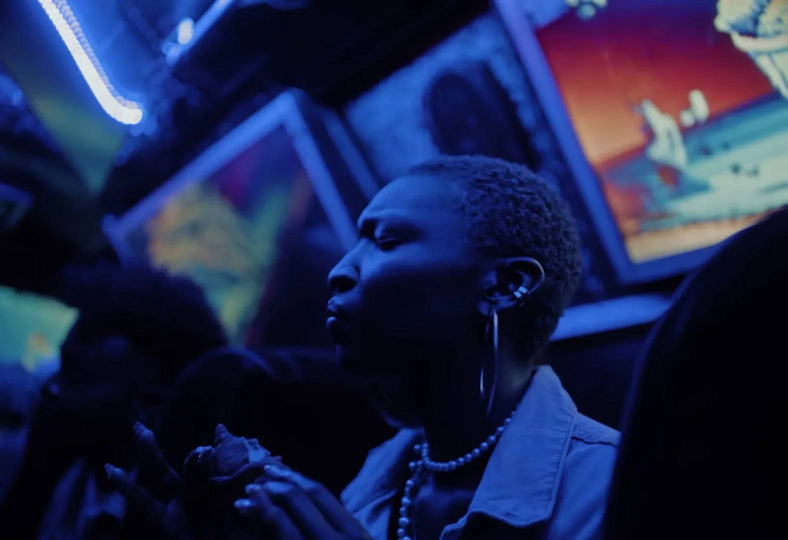 A person with short hair and pearl necklace eats a piece of chicken in a dark, blue-lit room with screens.