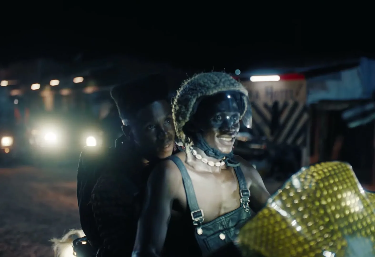 Two young men, one with a furry helmet and overalls, the other behind him, ride a motorcycle at night in a dimly lit, possibly rural, area.