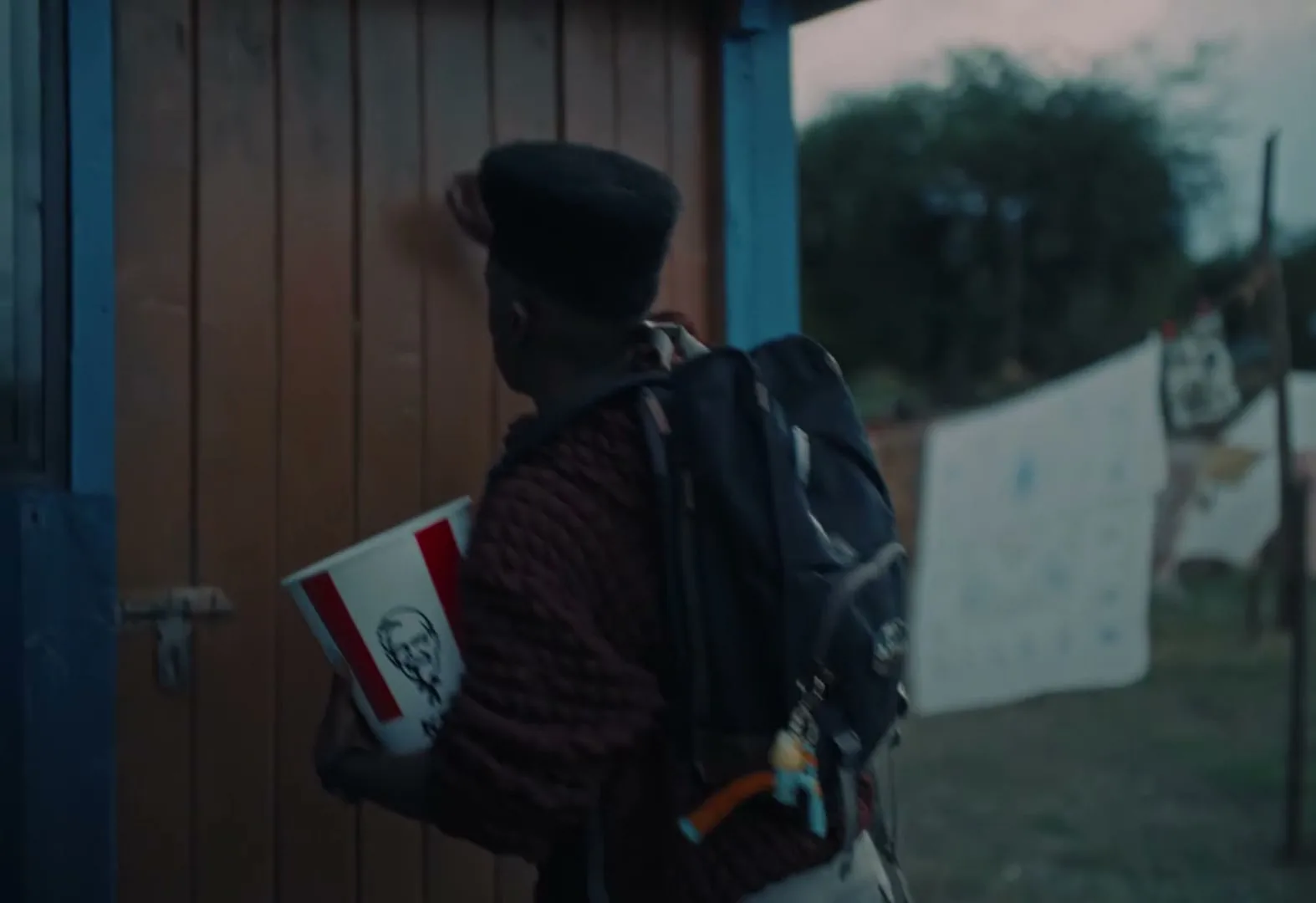 A young man with a backpack and a KFC bucket knocks on a wooden door in a rural outdoor setting at dusk.