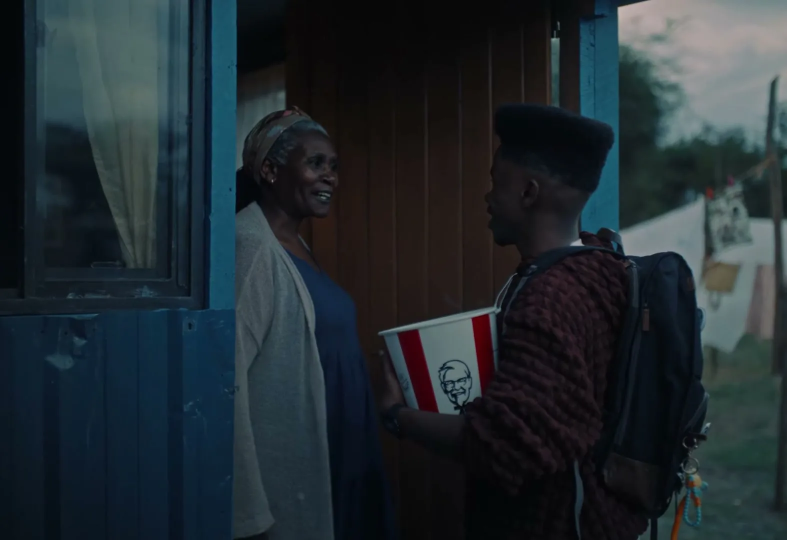 A young man with a backpack holds a KFC bucket, talking to an older woman in a doorway at dusk.