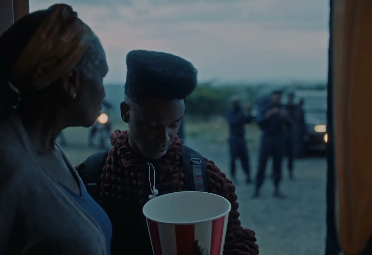 A young man with a unique haircut holds a KFC bucket and talks to an older woman at a doorway, with a group of people and vehicles in the background at dusk.
