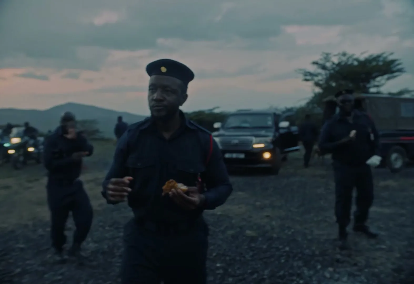 A male police officer in a dark uniform and cap holds a piece of fried chicken while standing outdoors with other officers and vehicles at dusk.