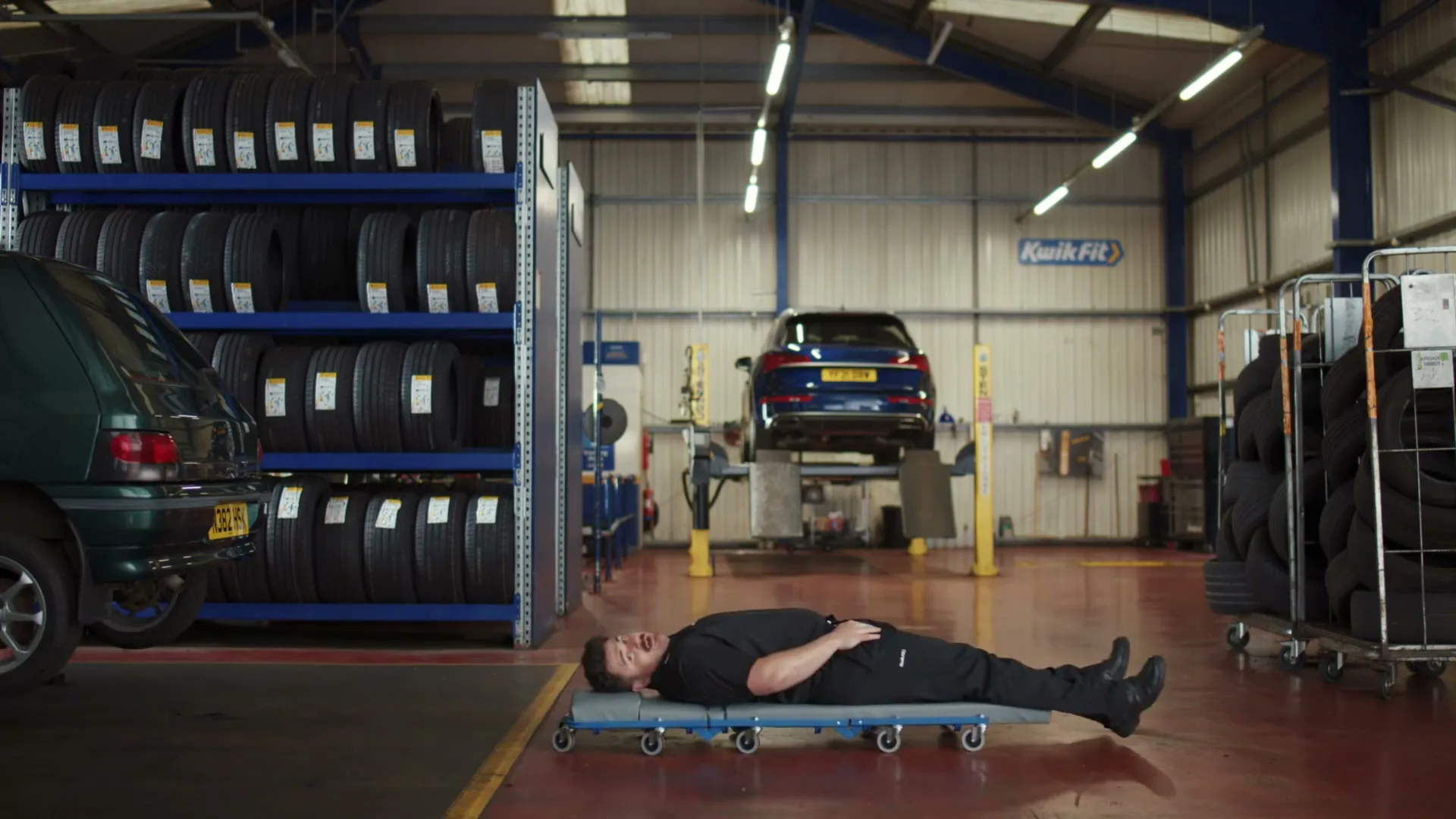 A male mechanic in a black uniform lies on a creeper, looking up and talking, in a Kwik Fit garage with cars and tires.