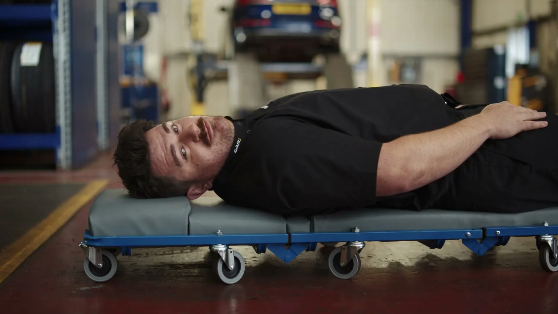 A male mechanic in a black uniform lies on a creeper, looking up at the camera in a car repair shop.