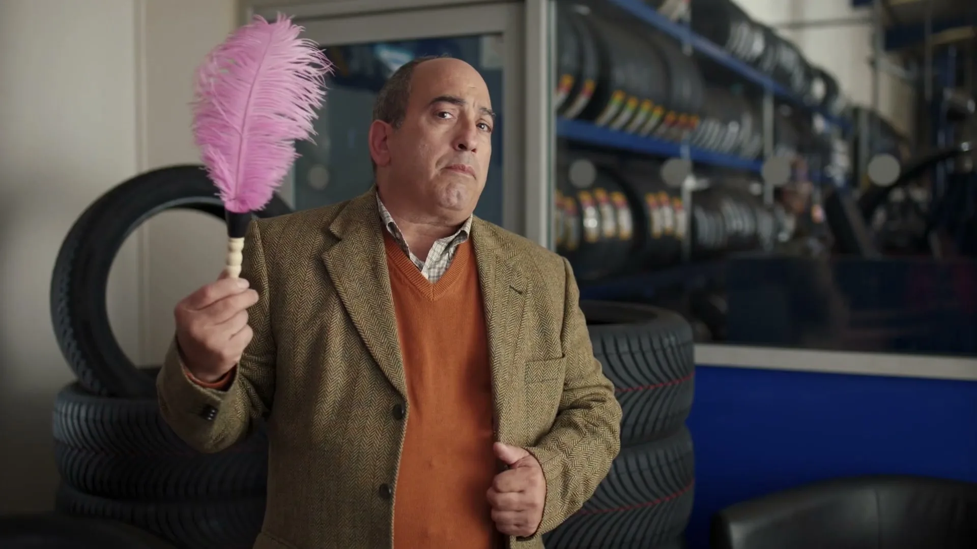 A man in a tweed jacket and orange sweater pulls a pink feather duster from his jacket in a tire shop.
