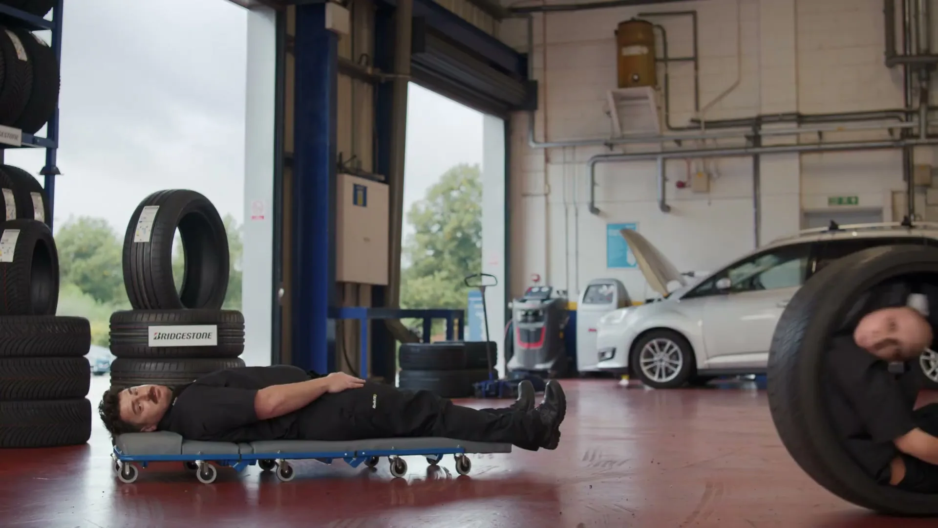 Two mechanics in black uniforms are in a garage, one lying on a creeper and the other inside a tire, as they move across the floor.