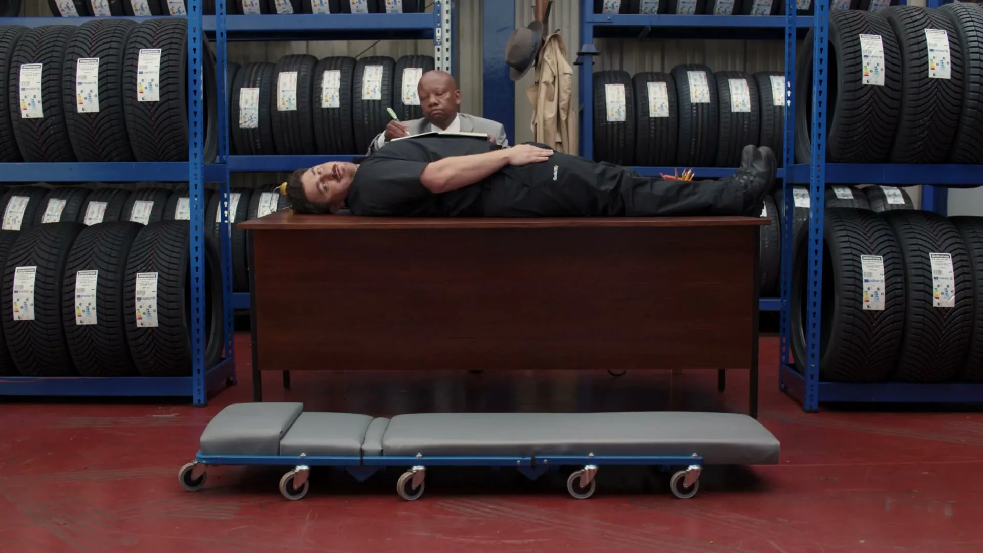 A man in a black uniform lies on a desk while another man in a suit sits behind him, taking notes in a tire shop.
