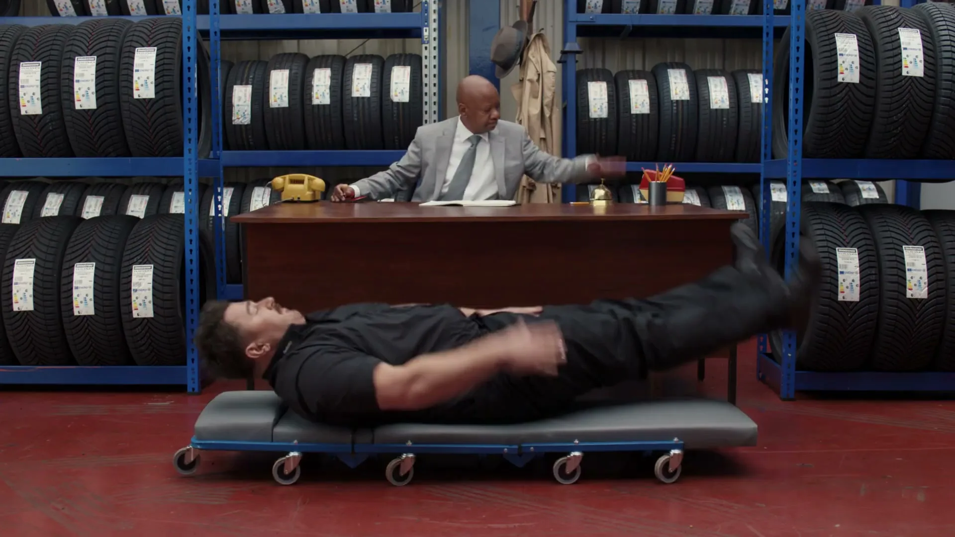 A man in a black uniform lies on a mechanic's creeper, being pushed by another man in a suit behind a desk, in a tire shop.