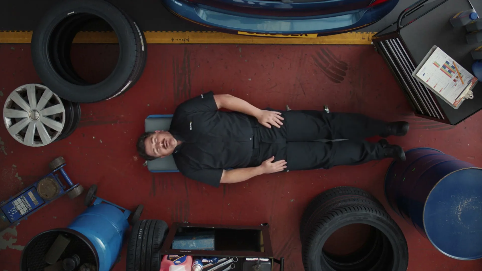A male mechanic in a black uniform lies on a creeper on a red garage floor, surrounded by tires and tools, looking up and talking.