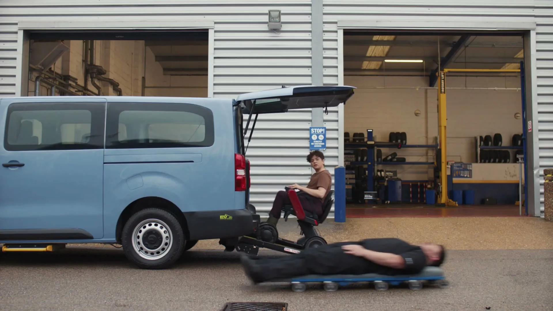 A young man in a brown shirt and dark pants sits in a mobility scooter, which is on a ramp extending from the back of a light blue van, as a mechanic on a creeper rolls past him at a garage.
