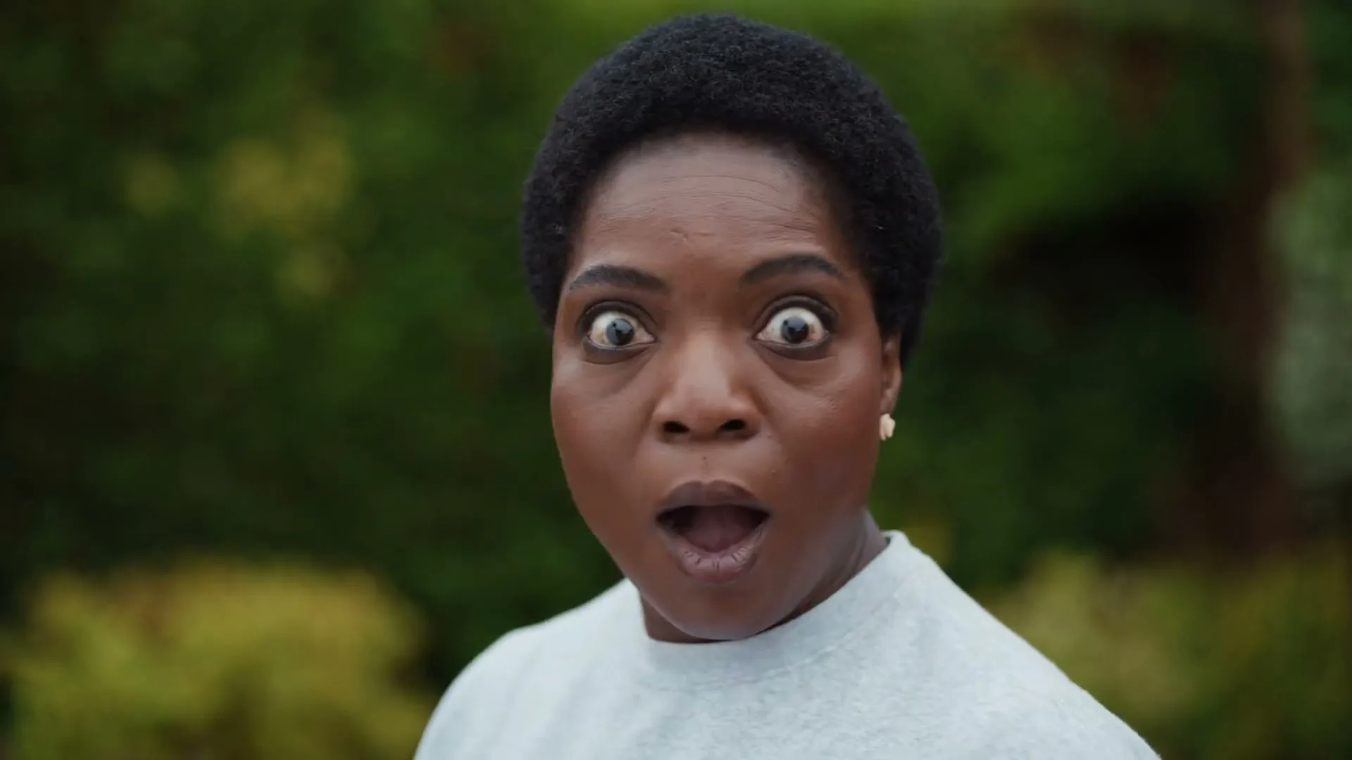 A Black woman with short, dark hair and a light grey sweatshirt looks surprised with wide eyes and an open mouth, outdoors against a green background.