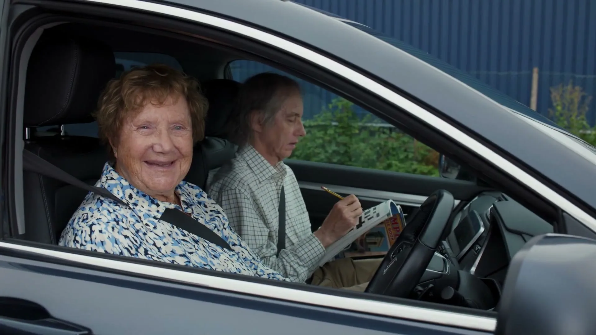An elderly woman with curly red hair smiles at the camera from the passenger seat of a car, while an elderly man in the driver's seat does a crossword puzzle.