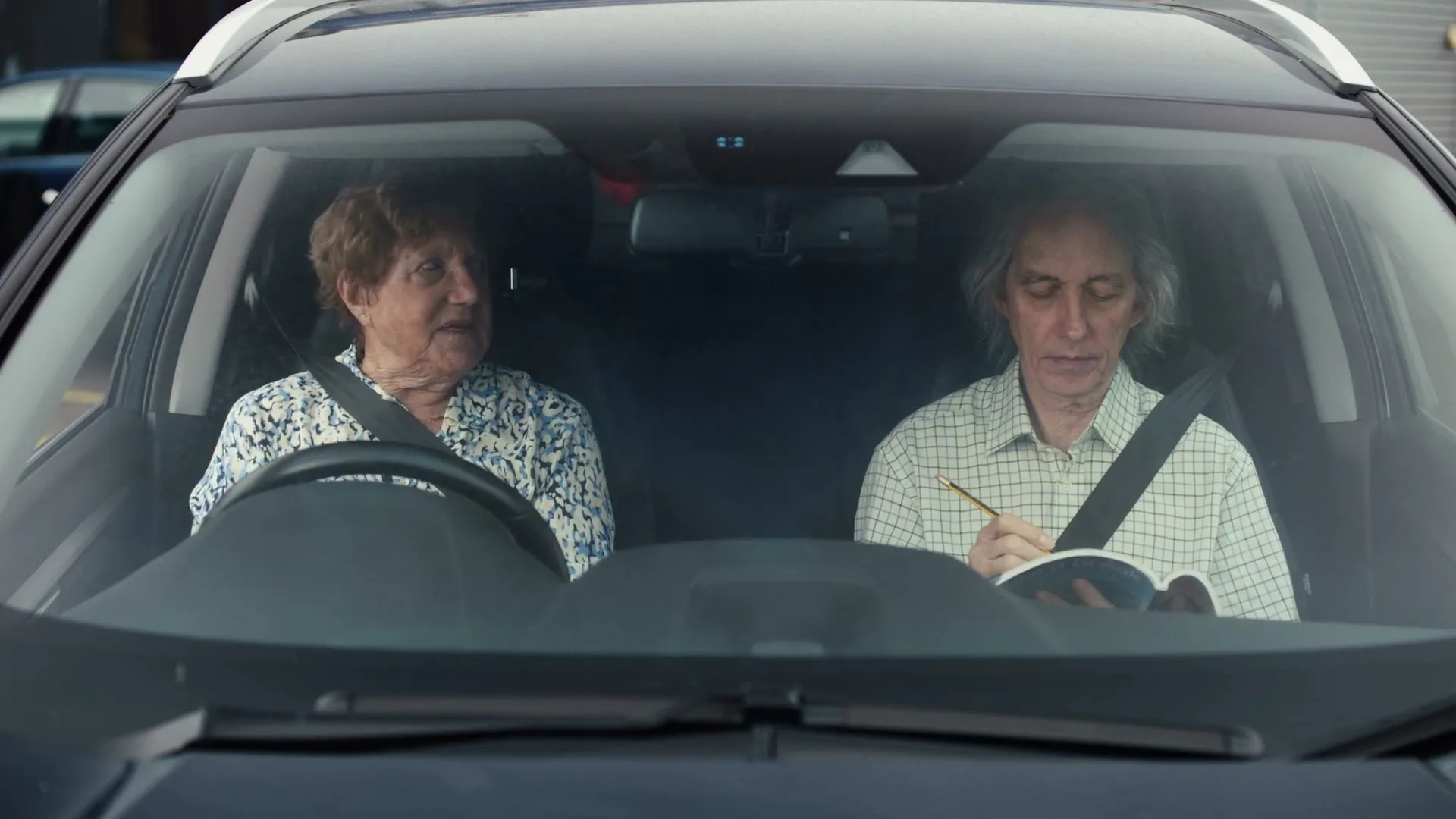 An elderly woman in a patterned top sits in the driver's seat of a car, looking at an elderly man in a plaid shirt who is writing in a book in the passenger seat.