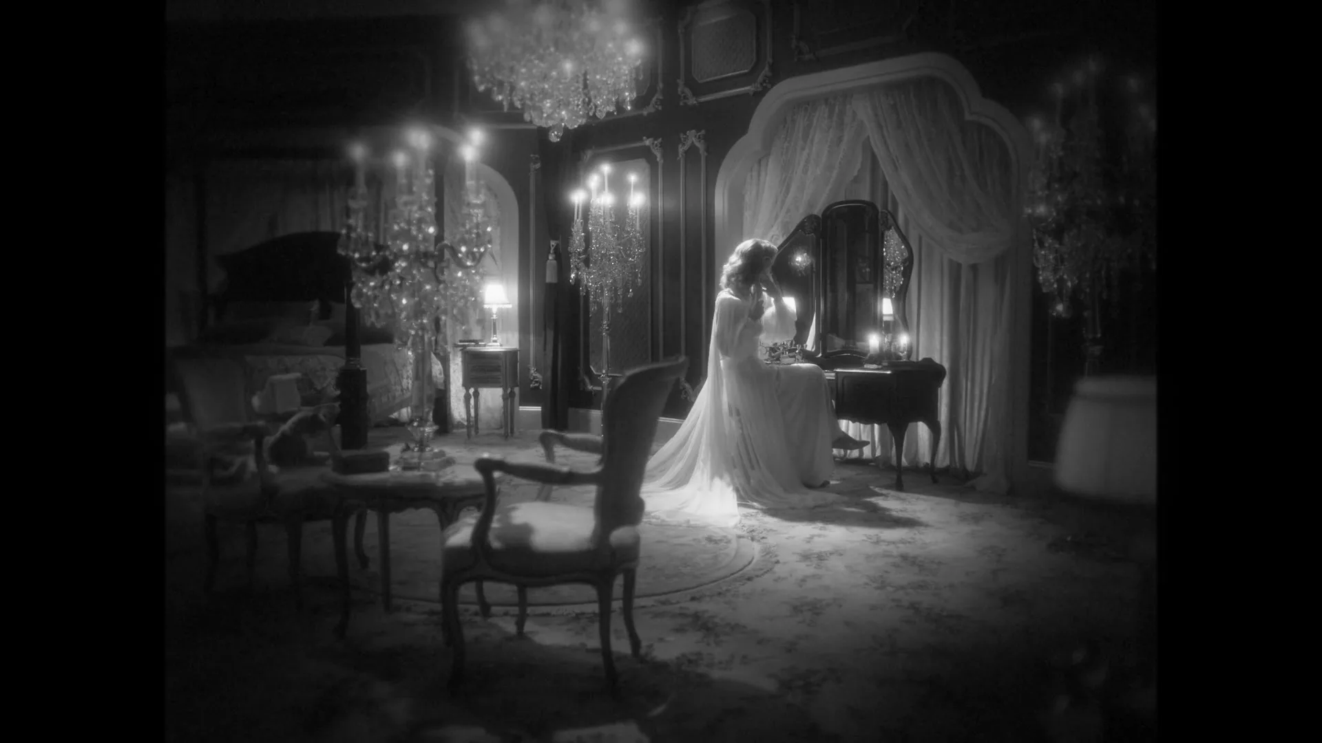A woman in a white nightgown sits at a vanity table in a dimly lit, opulent bedroom, looking at her reflection.