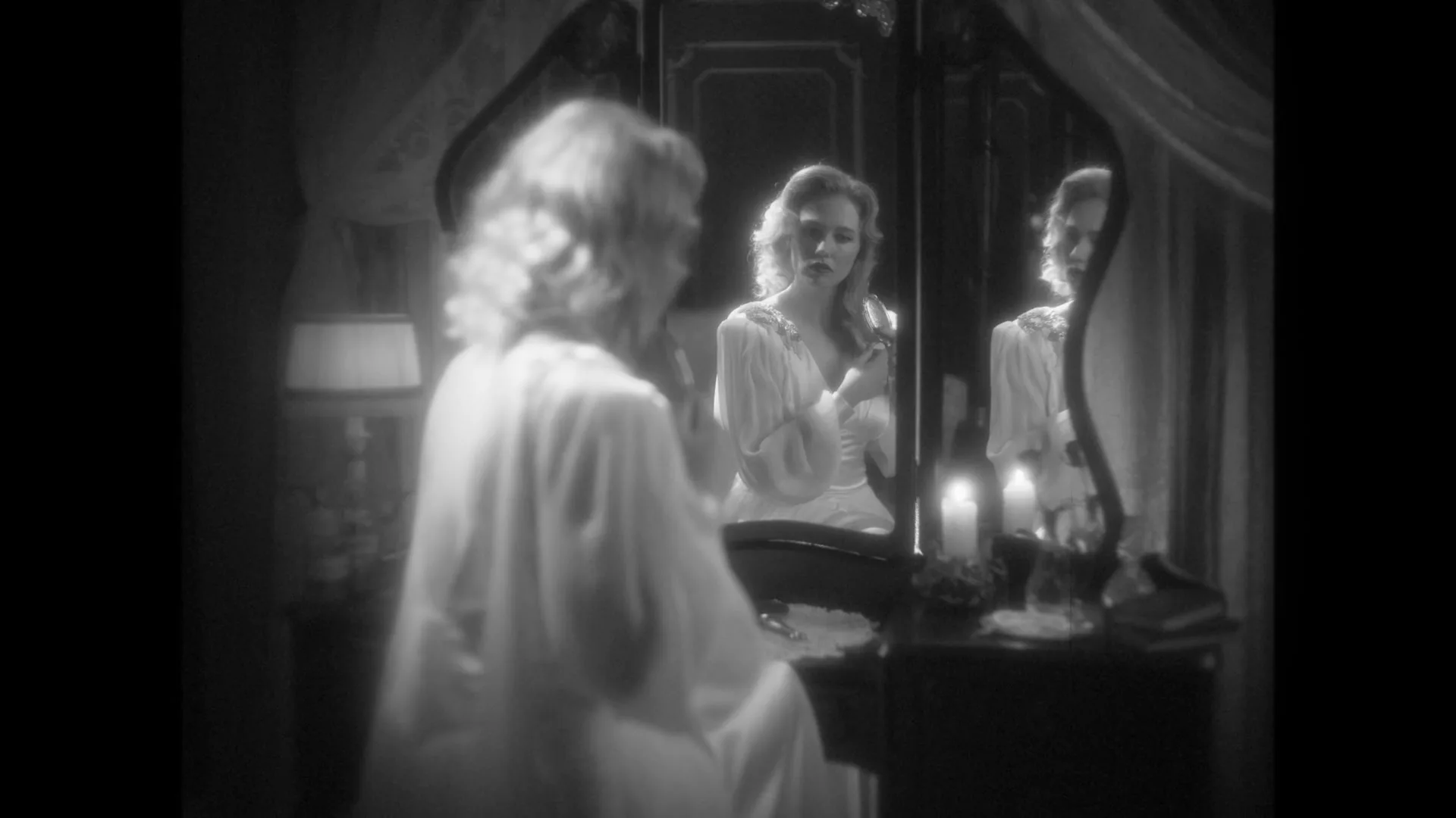 A woman in a white nightgown sits at a vanity, applying lipstick while looking at her reflection in a mirror in a dimly lit bedroom.