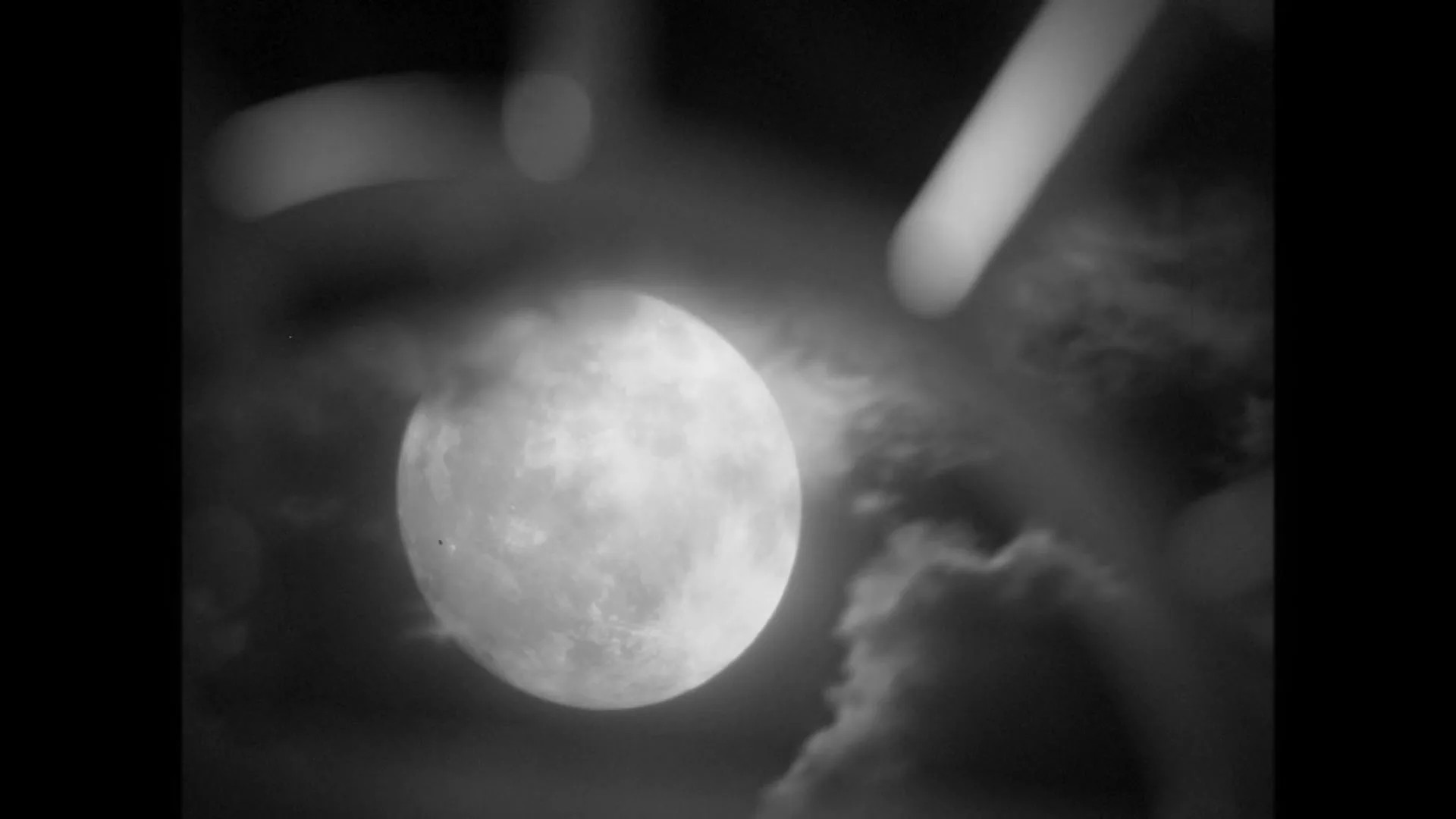 A full moon is seen through a blurry foreground element, with clouds in the night sky, in a black and white shot.