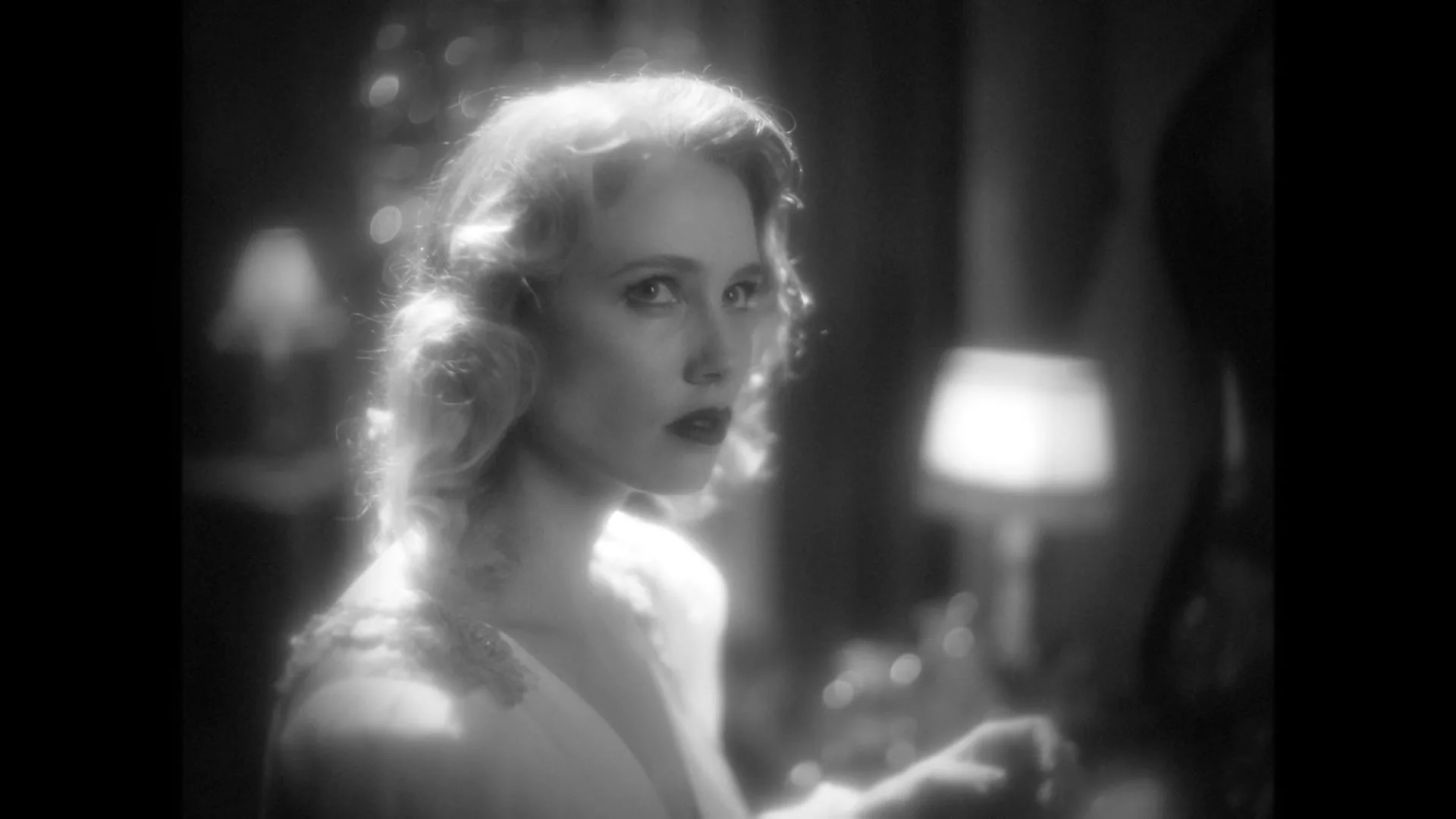 A woman with blonde hair and dark lipstick looks over her shoulder in a dimly lit room, in a black and white shot.