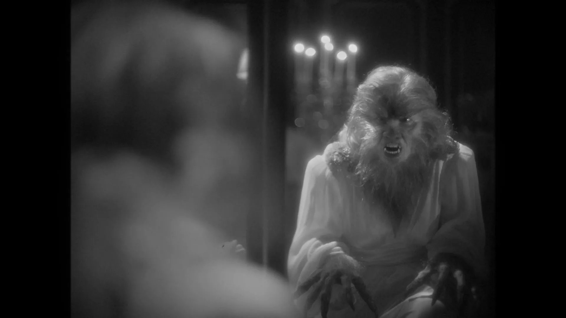 A black and white shot of a werewolf in a white robe, with glowing eyes and sharp teeth, looking menacingly at its reflection in a mirror.
