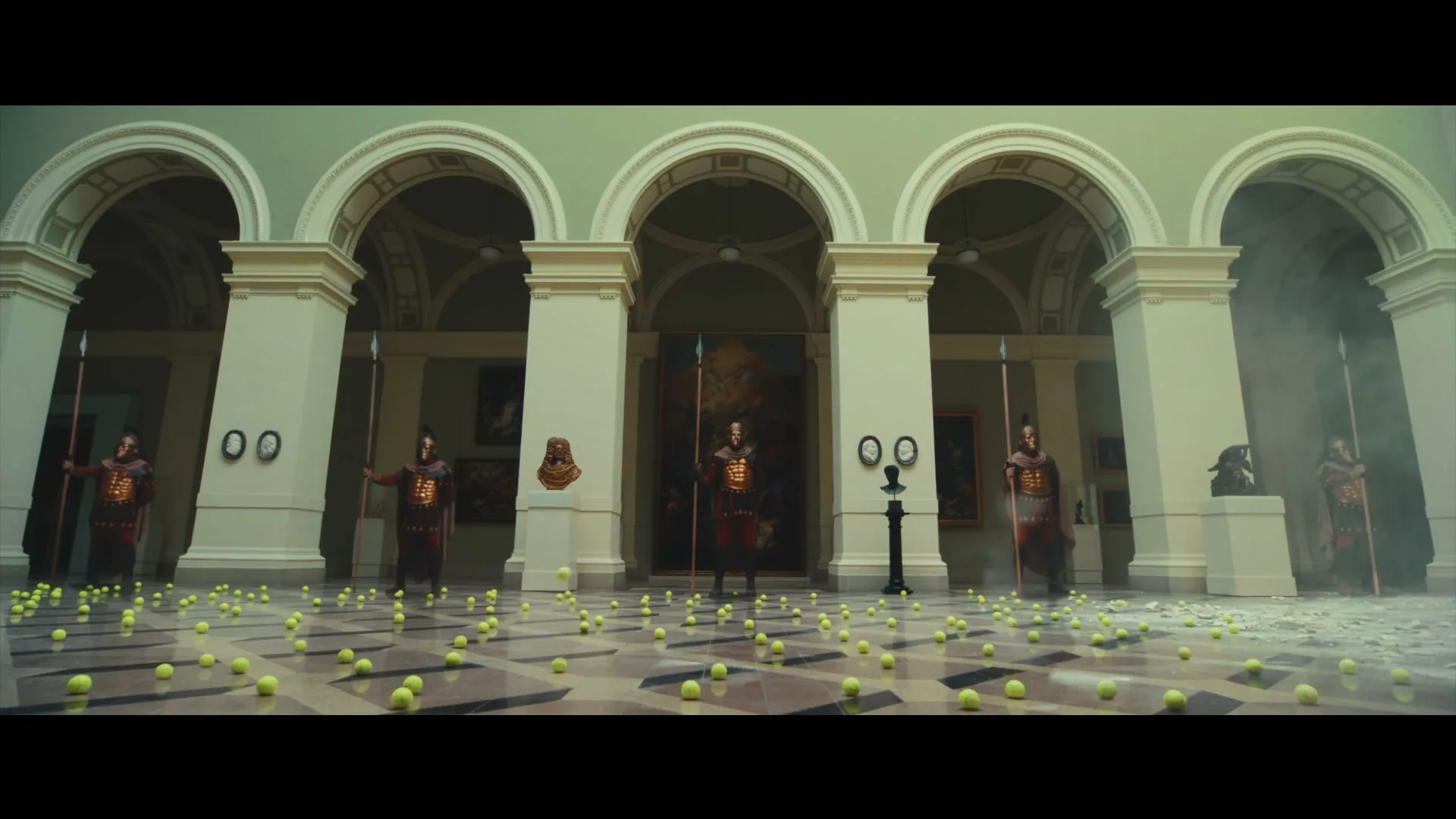 Five Roman guards stand in a grand hall with tennis balls scattered on the floor, as a tennis ball bounces in slow motion.
