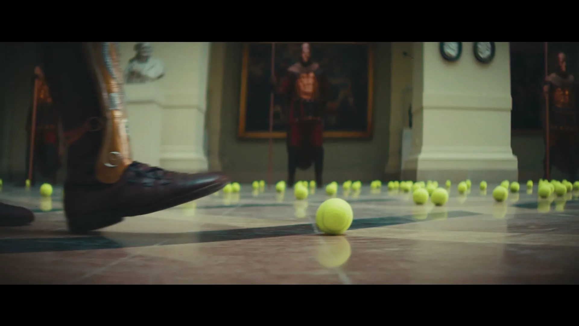A person in ancient warrior attire walks through a grand hall, stepping on a tennis ball among many scattered on the floor.