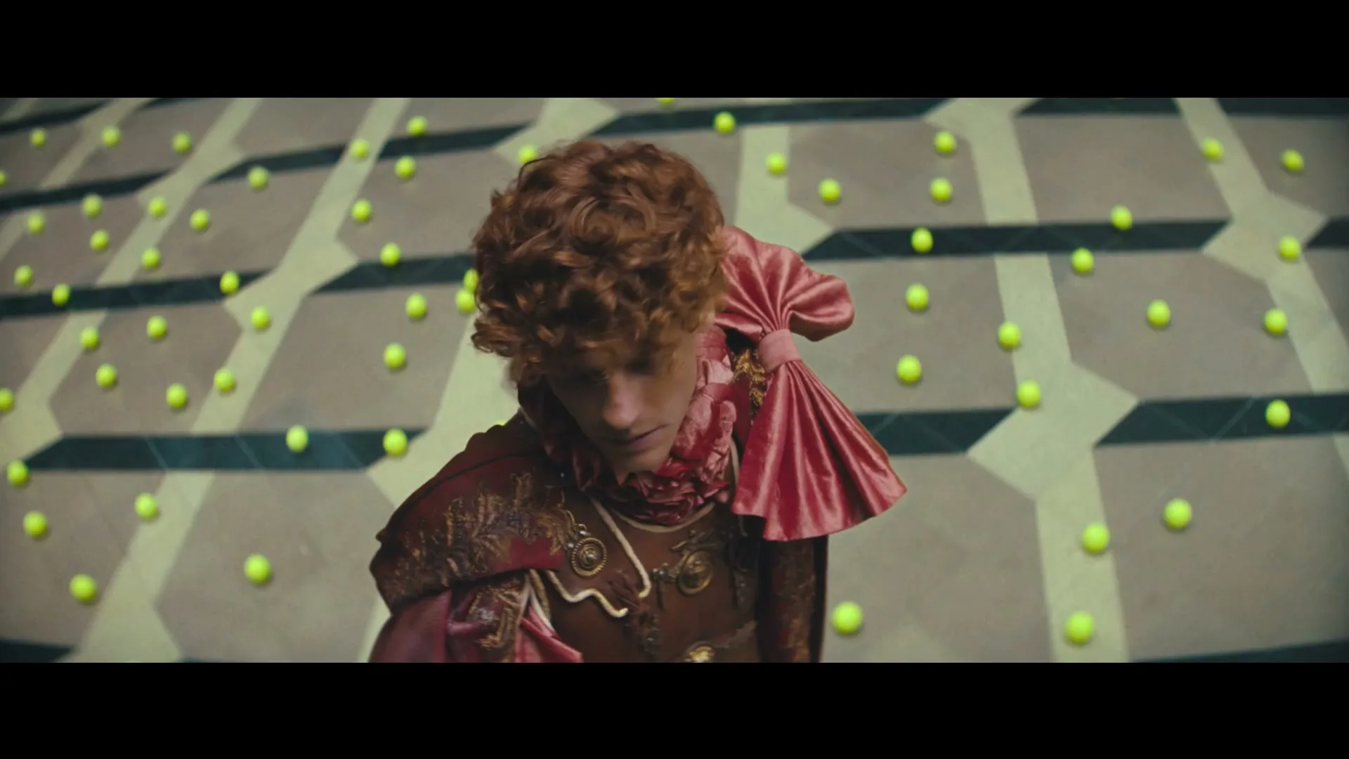 A young man with curly red hair and an elaborate pink and gold costume looks up, surrounded by a patterned floor with glowing green orbs.