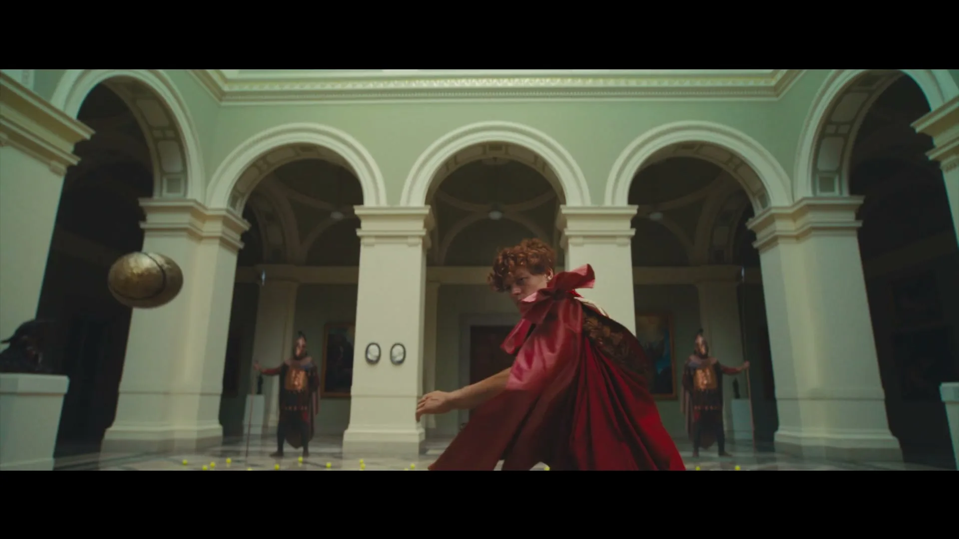 A young man in a red cape plays tennis with a golden orb in a grand hall, as two guards stand by.