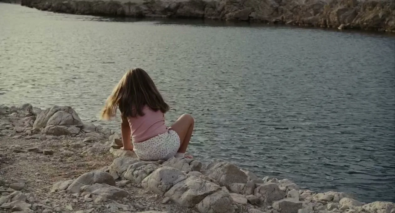 A young girl with long brown hair sits on rocks by the water, looking out at the calm sea during the day.