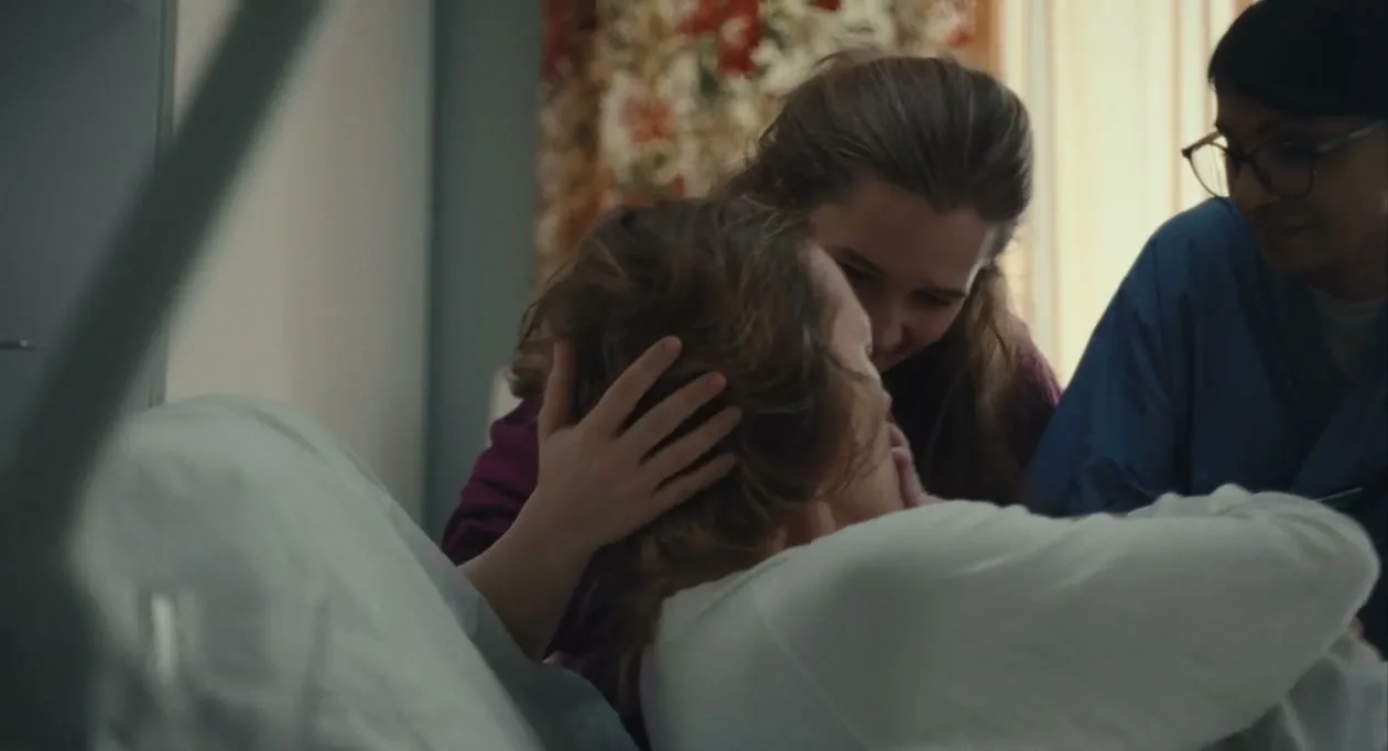 A woman in a hospital bed holds her newborn baby, while another woman and a nurse lean over them, smiling.