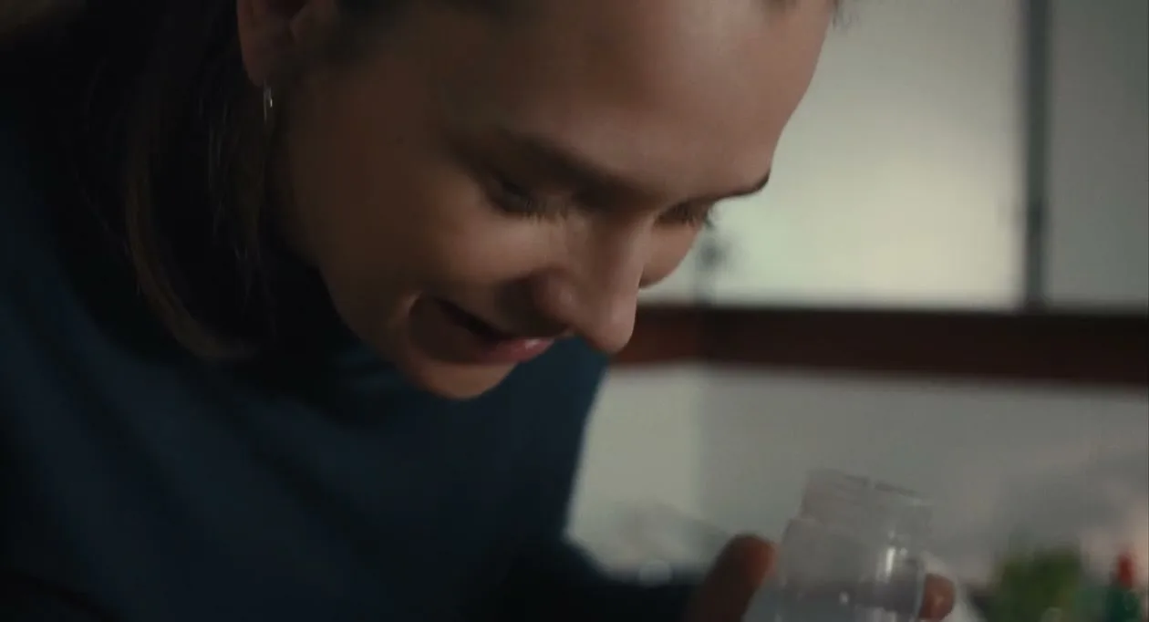 A young woman with brown hair in a ponytail, wearing a blue turtleneck, smiles while holding a small plastic container in a kitchen.