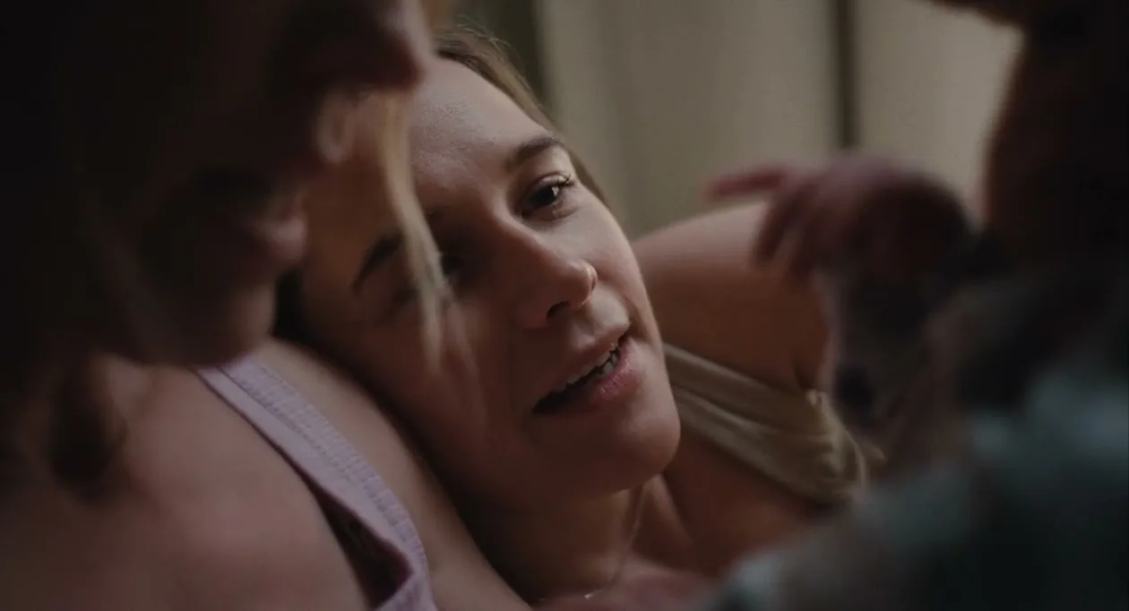 A young woman with light brown hair smiles softly while looking at a baby, as another person's head is visible in the foreground, in a dimly lit room.