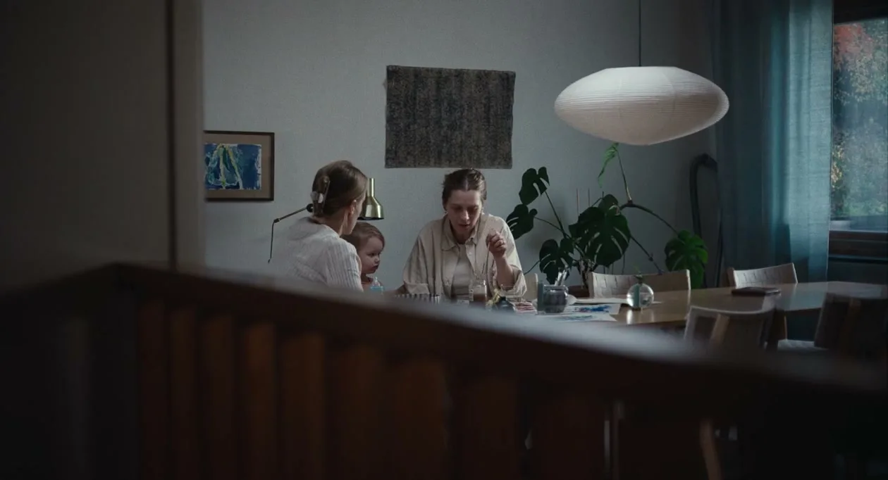 Two women and a baby sit at a dining table, seen through a blurred foreground, in a dimly lit interior.