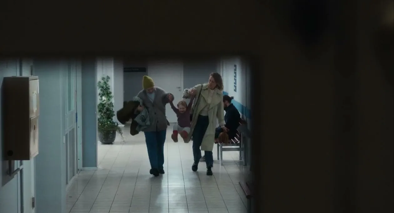A family, a woman in a yellow beanie, a woman in a trench coat, and a small child, walk down a hospital corridor, seen through a doorway.
